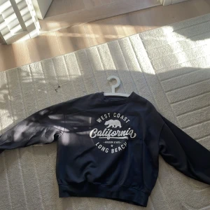 Svart sweatshirt med tryck - Säljer en svart sweatshirt med tryck 'West Coast California Long Beach' i vitt. Tröjan har långa ärmar och en rund halsringning. Perfekt för en avslappnad stil.