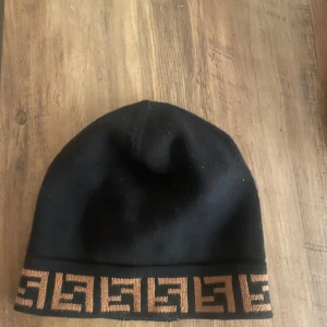 Svart mössa från Fendi - Snygg svart mössa från Fendi med ett brunt geometriskt mönster längs kanten. Perfekt för att hålla värmen med stil. Märkesetikett på insidan.