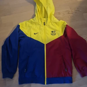 Flerfärgad vindjacka från Nike - Säljer en cool flerfärgad vindjacka från Nike med FCB-logga. Jackan har en huva och dragkedja framtill. Färgerna är gul, blå och röd, vilket ger en sportig look. Perfekt för fotbollsfans! 💙❤️💛