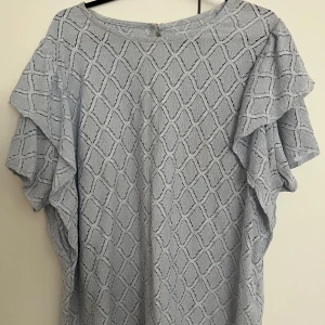 Ljusblå blus med volangdetaljer - Säljer en ljusblå blus med ett elegant mönster och volangdetaljer längs ärmarna. Perfekt för en stilren look. Den är i Xl men passar S/M också