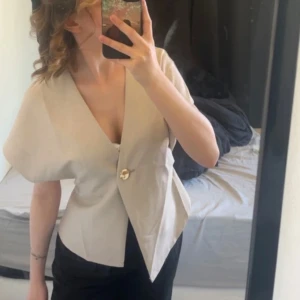 Beige topp med gulddetalj  - Säljer en stilren beige topp med en knappdetalj framtill. Blusen har en v-ringad design och korta ärmar. Köpt här på Plick, oanvänd. Storlek S!🩷
