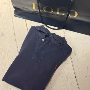 Mörkblå pikétröja från Polo - Säljer en klassisk mörkblå pikétröja från Polo med korta ärmar och en liten röd logga på bröstet. Perfekt för en stilren och avslappnad look. Tröjan har en knappslå med två knappar och en traditionell krage.