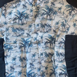 Hawaiiskjorta med palmmönster - Snygg kortärmad hawaiiskjorta med ett tropiskt palmmönster i blått och vitt. Perfekt för sommarens äventyr och strandhäng. Skjortan har knappar framtill och en avslappnad passform.