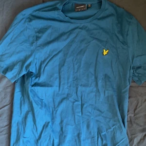 Blå t-shirt från Lyle & Scott - Säljer en blå t-shirt från Lyle & Scott med korta ärmar och en gul broderad logga på bröstet. Perfekt för en avslappnad stil.