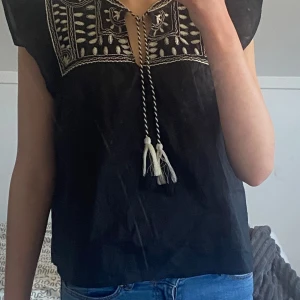 Svart blus med broderi och knytband - Säljer en svart blus med vackert vitt broderi på bröstet och knytband med tofsar framtill. Blusen har korta ärmar och en lös passform, perfekt för en bohemisk stil.