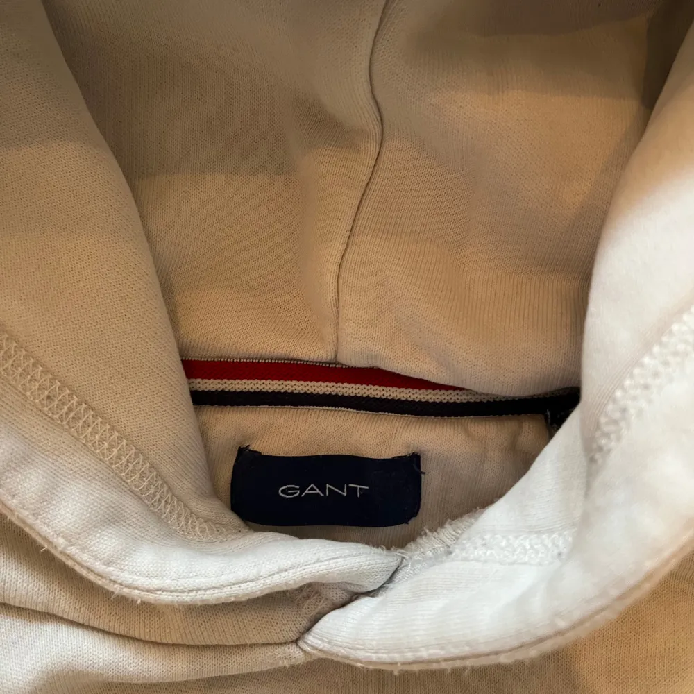 Säljer en stilren vit hoodie från GANT med broderad logga och emblem på bröstet. Tröjan har en klassisk känguruficka och en bekväm huva. Perfekt för en avslappnad stil.. Neuletakit & Villapaidat.