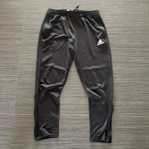 Svarta träningsbyxor från Adidas - Säljer ett par svarta träningsbyxor från Adidas med elastisk midja och dragkedjor vid bensluten. Byxorna har en vit Adidas-logga på ena benet och är perfekta för sport och träning.