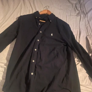 Svart långärmad skjorta med broderad logga - Snygg svart långärmad skjorta från Ralph Lauren med klassisk krage och knappar framtill. Skjortan har en broderad logga på bröstet som ger en stilren touch. Perfekt för en elegant look.