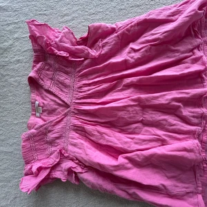 Rosa blus från Gina Tricot - Säljer en söt rosa blus från Gina Tricot med volangdetaljer vid ärmarna. Blusen har en lös passform och är perfekt för varmare dagar. Den har en smockad design vid axlarna och runt halsen.