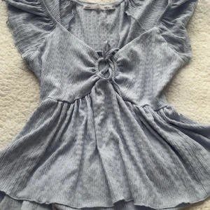 Ljusblå topp från 157 - Säljer en söt ljusblå topp från 157 med korta ärmar och knytning framtill. Toppen har en luftig passform och är perfekt för varma dagar. Den har en lätt ribbad struktur som ger en fin detalj.