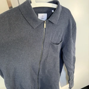  stickad tröja med dragkedja från Bläck - Säljer en stilren stickad tröja från Bläck med dragkedja framtill och en bröstficka. Perfekt för en avslappnad look. Tröjan har en klassisk krage och långa ärmar.