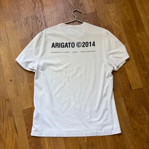 KrämVit t-shirt från Arigato - Säljer en stilren krämvit t-shirt från Arigato med tryck både fram och bak. Perfekt för en avslappnad look. T-shirten har korta ärmar och en klassisk passform. 