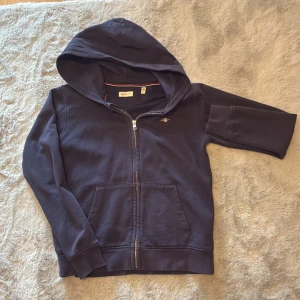 Mörkblå hoodie från GANT - Säljer en snygg mörkblå hoodie från GANT med dragkedja och huva. Perfekt för kyliga dagar. Den har praktiska fickor framtill och en stilren design. Passar ungdomar i åldern 13-14 år.