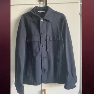Snygg mörkblå overshirt från Acne Studios med knappar framtill och två stora bröstfickor. Perfekt för lager-på-lager-stil. Klassisk krage och långa ärmar ger en stilren look.