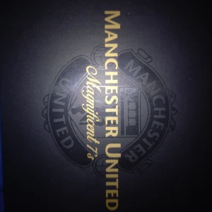 Manchester United Magnificent 7's Samlarbox - Exklusiv samlarbox med Manchester United-tema, innehållande miniatyrtröjor av legendariska spelare som Best, Robson, Cantona, Beckham och Ronaldo. Varje tröja har nummer 7 och boxen pryds av klubbens emblem.