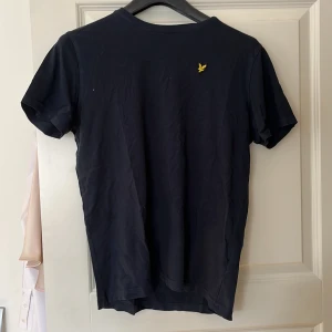 Mörkblå t-shirt från Lyle & Scott - Säljer en stilren blå t-shirt från Lyle & Scott med korta ärmar. Den har en gul broderad logga på bröstet. Perfekt för en avslappnad look.