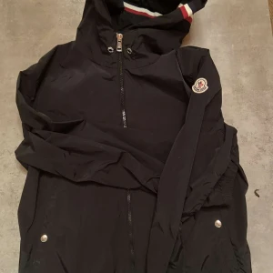 Moncler windbreaker  - Säljer en stilren svart vindjacka från Moncler med dragkedja och huva. Jackan har Monclers logotyp på ärmen och ribbade detaljer vid ärmslut. Perfekt för blåsiga dagar.