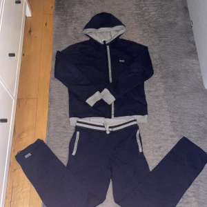 Mörkblå hoodie från Hugo Boss - Säljer en snygg mörkblå hoodie från Hugo Boss med gråa detaljer. Tröjan har en dragkedja framtill och en bekväm huva. Perfekt för en avslappnad stil. Knappt använd.