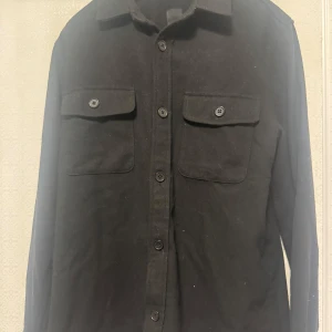 Svart långärmad skjorta med knappar - Snygg svart långärmad skjorta med knappar framtill och två bröstfickor med knäppning. Perfekt för en stilren look.