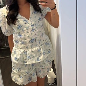 Blommig vit klänning med volanger - Vit klänning med blå blommönster och volangdetaljer. Klänningen har korta ärmar och en v-ringad halslinje med spetskant. Perfekt för sommarens varma dagar. HELT NY, använd EN GÅNG. Priser går alltid att diskutera