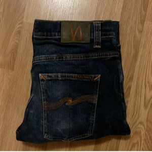 Mörkblå jeans från Nudie - Snygga mörkblå jeans från Nudie med klassisk femficksdesign och orange sömmar. Jeansen har en knappgylf och en stilren passform som passar perfekt till vardags. Perfekt för dig som gillar en tidlös stil.