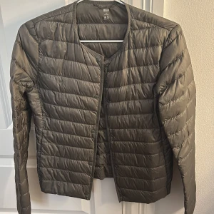 Grön lättviktsjacka från Uniqlo - Säljer en grön lättviktsjacka från Uniqlo i storlek XS. Jackan är quiltad och har en stilren design utan krage. Perfekt för övergångsperioder med sin vattenavvisande funktion. Långärmad och bekväm att bära.