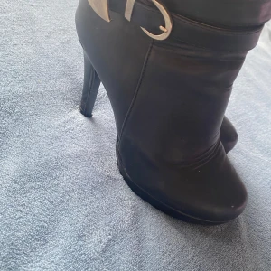 Svarta stövletter med klack - Snygga svarta stövletter i skinn med klack och rund tå. De har en dekorativ rem med spänne runt ankeln och dragkedja på sidan för enkel på- och avtagning.