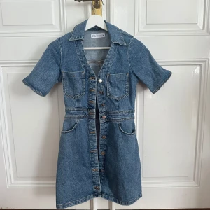 Jeansklänning från Zara - Snygg jeansklänning från Zara med knappar framtill och korta ärmar. Klänningen har en klassisk krage och två bröstfickor. Perfekt för en avslappnad stil. Skulle säga att den är en mindre xs.
