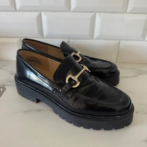 Svarta loafers i skinn med grov sula - Snygga svarta loafers i äkta skinn med chunky, räfflad sula och dekorativt guldfärgat spänne över foten. Klassisk design med rund tå och platt sula, perfekta för en trendig vardagslook. Knappt använda!