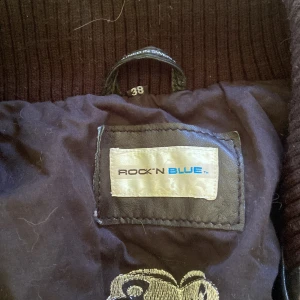 Svart skinnjacka från Rock'n Blue - Säljer en svart skinnjacka från Rock'n Blue med stora ribbade muddar vid krage, ärmslut och nederkant. Jackan har flera fickor med lock och dragkedja samt dekorativa knappar. Perfekt för dig som vill ha en cool och tidlös stil.