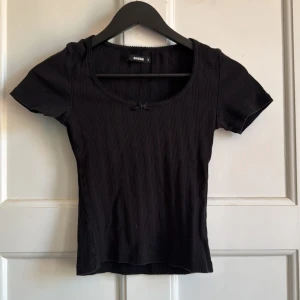 Svart ribbad topp från BikBok - Säljer en svart ribbad topp från BikBok med korta ärmar och rundad urringning. Toppen har en liten rosett framtill och är figurnära i modellen. Perfekt att matcha med jeans eller kjol.