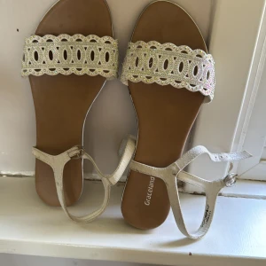 Vita sandaler med glittrig spets från Graceland - Säljer ett par vita sandaler från Graceland med bred rem i glittrig spets och dekorativa detaljer. Skorna har öppen tå och justerbar rem runt ankeln. Perfekta för sommaren och ger en snygg touch till din outfit.