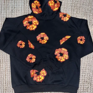 Svart hoodie med röda och gula blommor från Denim Tears - Säljer en svart hoodie från Denim Tears med unika röda och gula blommor tryckta över hela tröjan. Hoodien har en klassisk passform, huva och magficka. Perfekt statement-plagg för dig som vill sticka ut.