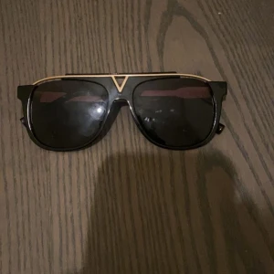 Svarta solglasögon med gulddetaljer - Snygga svarta solglasögon med markerad gulddetalj upptill och vid näsryggen. Rektangulär form med bred båge och mörka glas. Perfekt accessoar för att lyfta din stil.