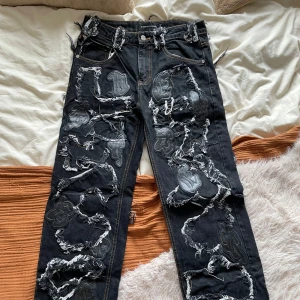 Svarta jeansbyxor med patchwork från Racer Worldwide - Unika svarta jeans från Racer Worldwide med råa, slitna detaljer och stora svarta patchar i olika former. Byxorna har normal passform och raka ben, med coola kontrastsömmar och en edgy streetwear-vibe.