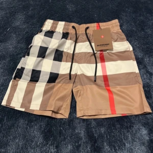 Burberry badshorts med klassiskt rutmönster - Snygga badshorts från Burberry med det ikoniska rutmönstret i beige, svart, vitt och rött. Shortsen har elastisk midja med svart dragsko och innerbyxa i mesh. Perfekta för sommardagar vid stranden eller poolen.