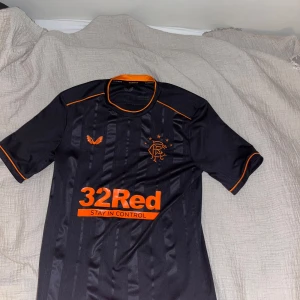 Rangers FC svart fotbollströja med orange detaljer - Säljer en svart Rangers FC fotbollströja från Castore med orange logga, detaljer och sponsortryck. Tröjan har korta ärmar, rund hals med orange insida och tunna vertikala ränder på framsidan. Perfekt för dig som gillar snygga matchtröjor!