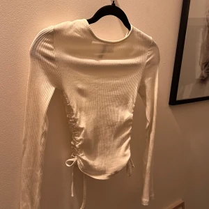 Vit ribbad långärmad topp med knyt - Säljer en vit långärmad topp med ribbad struktur och knyt i sidan som ger en snygg rynkad effekt. 💞