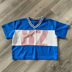 Blå croppad t-shirt från Better Bodies - Säljer en blå croppad t-shirt från Better Bodies med vit och röd detalj framtill och stor siffra 82. T-shirten har v-ringning med randig kant och korta ärmar. Perfekt för en sportig look.