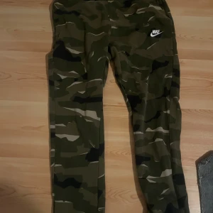 Nike camouflage träningsbyxor - Säljer ett par Nike träningsbyxor i camouflage-mönster. Byxorna har en smal passform och är perfekta för träning. De är gröna och har Nike-loggan tryckt på ena benet.