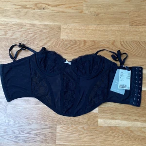 Svart spetskorsett från H&M - Säljer en svart korsett från H&M med spetsdetaljer och justerbara axelband. Korsetten har flera hyskor i ryggen för perfekt passform och är tillverkad i ett genomskinligt syntetmaterial med vacker spets framtill.