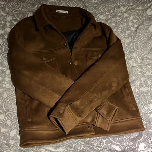 Vår jacka  - Snygg brun mockajacka från Zara med klassisk krage och knappar framtill. Jackan har två bröstfickor med lock och knappar samt slät baksida. Fodrad med svart tyg för extra komfort. Perfekt för dig som gillar stilren och tidlös look.