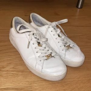 Vita sneakers från Michael Kors med gulddetaljer - Snygga vita sneakers från Michael Kors med guldfärgade detaljer på häl och snörning. Skorna har rund tå, platt sula och är tillverkade i skinn. 
