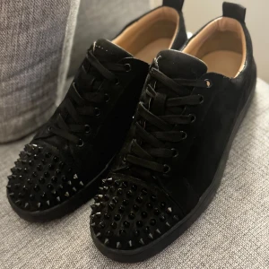 Svarta sneakers med nitar och röd sula - Säljer ett par svarta sneakers från Christian Louboutin med svarta nitar på tån och den ikoniska röda sulan. Skorna har snörning och är tillverkade i mocka med läderdetaljer. Perfekta för dig som vill sticka ut med en edgy stil.