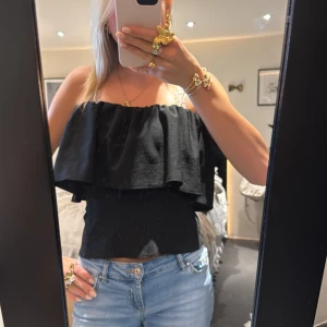 Svart offshoulder topp med volang - Säljer en svart offshoulder topp med bred volang runt hela överdelen. Toppen är croppad och har en snygg, avslappnad passform. Perfekt att matcha med jeans eller kjol för en trendig look.