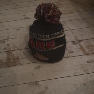 Svart och röd Åre-mössa med tofs - Mysig svart stickad mössa från Åre med stor röd, vit och svart tofs på toppen. Texten 'SWEDEN' och 'ÅRE' i vitt och rött samt bergmotiv framtill. Perfekt för kalla dagar.