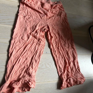 Persikofärgade byxor med volang nertill från Name It( är till barn) - Säljer ett par persikofärgade byxor från Name It i mjukt material. Byxorna har volangdetalj vid benslut och är croppade i modellen. Perfekta för vår och sommar! Storleken 140/cr 10 år 