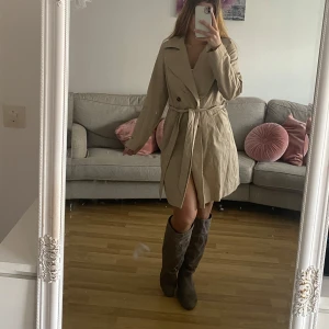 Beige trenchcoat med bälte - Säljer en klassisk beige trenchcoat med bälte i midjan och stora slag. Jackan har dubbelknäppning med dekorativa knappar framtill och långa ärmar. Perfekt för vår och höst, ger en stilren och tidlös look.
