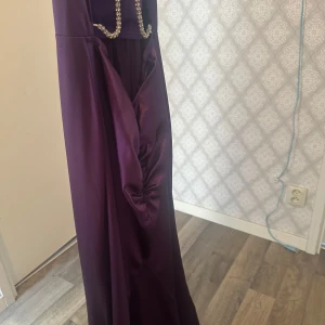Lila långklänning med strassdetaljer - Säljer en elegant lila långklänning med halterneck och dekorativa strassband över ryggen. Klänningen har en åtsittande passform upptill och ett vackert fall. Perfekt för bal eller festkvällar!