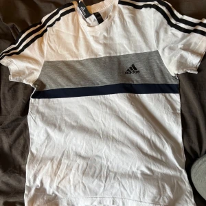 Vit och grå t-shirt från Adidas - Säljer en fräsch t-shirt från Adidas i vitt med grått och mörkblått band över bröstet. Klassiska svarta ränder på axlarna och Adidas-logga på bröstet. Perfekt för en sportig look.